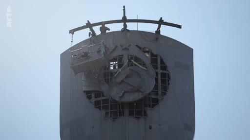 Des ouvriers utilisent une nacelle pour démonter l'emblème soviétique de la faucille et du marteau situé au sommet d'un monument imposant. Cette opération symbolique marque la décommunisation et le retrait des vestiges de l'ère soviétique.