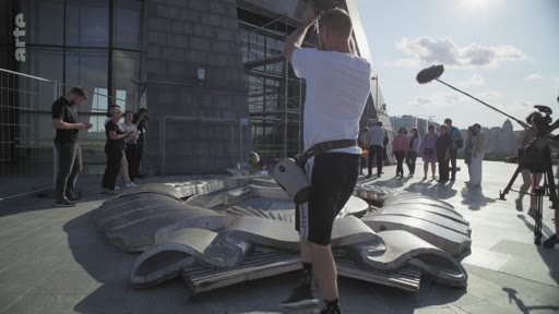Une équipe de tournage, incluant un cadreur avec une caméra et un preneur de son, filme un monument en forme d'étoile sur une place publique en extérieur. Le tournage se déroule sous un ciel ensoleillé avec plusieurs personnes en arrière-plan.