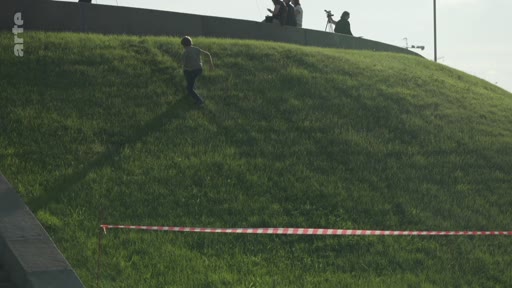 Un enfant court sur une colline herbeuse, probablement au pied d'un monument ou d'un site commémoratif, sous le regard de personnes assises en haut de la pente. Une bande de signalisation rouge et blanche est visible au premier plan.