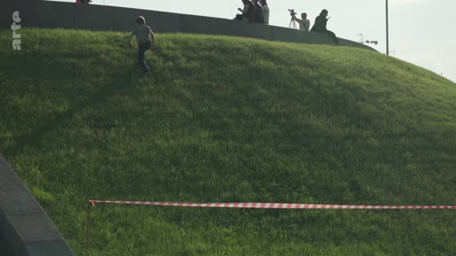 Un enfant monte à pied sur une colline recouverte d'herbe, délimitée par une rubalise rouge et blanche, avec des personnes assises en arrière-plan sur un muret. La scène se déroule en extérieur dans un espace public aménagé.