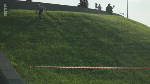 Un enfant descend en courant une colline recouverte d'herbe, délimitée par une rubalise, près d'un monument où se trouvent des spectateurs. La scène se déroule en extérieur dans un espace public.