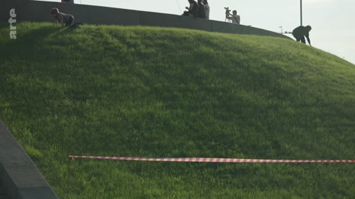 Des personnes, dont un caméraman, se trouvent sur une colline herbeuse délimitée par une rubalise, près d'un monument urbain.