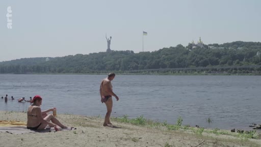 Vue sur une plage au bord du fleuve Dniepr à Kiev, en Ukraine, où des personnes se détendent. En arrière-plan, on distingue la statue monumentale de la Mère-Patrie et le monastère de la laure des Grottes de Kiev.