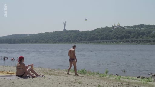 Vue estivale sur les rives du fleuve Dniepr à Kiev, en Ukraine, montrant des personnes se relaxant sur la plage. En arrière-plan, on distingue la statue monumentale de la Mère-Patrie et le drapeau ukrainien flottant au-dessus des collines boisées.