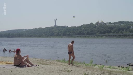Vue sur une plage au bord du fleuve Dniepr à Kiev, en Ukraine, où des personnes se détendent. En arrière-plan, on distingue la statue monumentale de la Mère-Patrie et les dômes de la laure des Grottes de Kiev.