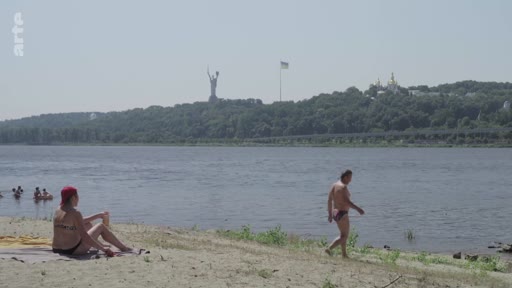 Vue d'ensemble d'une plage sur les rives du fleuve Dniepr à Kiev, en Ukraine, où des personnes se détendent. En arrière-plan, on distingue la statue monumentale de la Mère-Patrie et le monastère de la Laure des Grottes de Kiev.