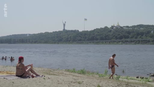 Vue panoramique d'une plage sur les rives du fleuve Dniepr à Kiev, en Ukraine, avec la statue monumentale de la Mère-Patrie et le monastère des Grottes de Kiev visibles sur la colline en arrière-plan. Des personnes se détendent au bord de l'eau par une journée ensoleillée.