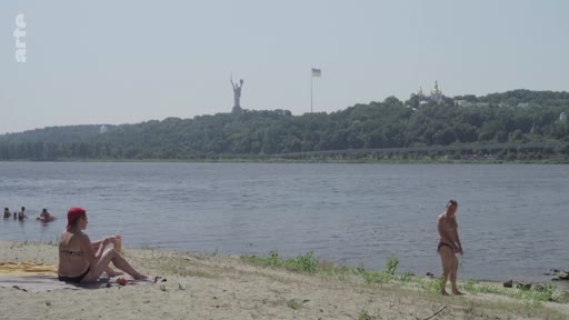 Vue panoramique d'une plage sur les rives du fleuve Dniepr à Kiev, en Ukraine, où des personnes se détendent. En arrière-plan, on distingue la statue monumentale de la Mère-Patrie et les dômes de la laure des Grottes de Kiev.