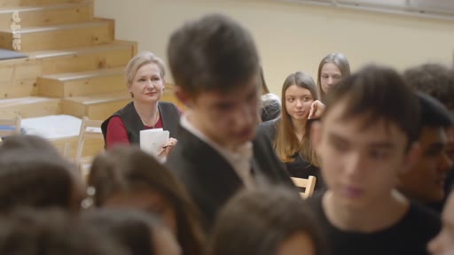 Une enseignante, assise en arrière-plan, observe attentivement ses élèves qui participent à une discussion ou un débat dans une salle de classe. L'ambiance est studieuse et concentrée, avec plusieurs jeunes étudiants au premier plan.