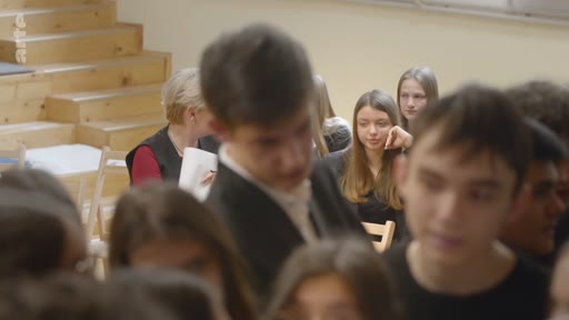 Un groupe d'élèves est réuni dans une salle de classe pour une discussion ou un échange, avec une enseignante visible en arrière-plan.