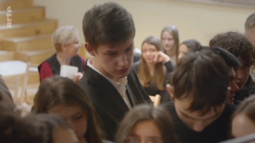 Un groupe d'élèves, dont un jeune homme au premier plan, est réuni dans une salle de classe pour une discussion ou un atelier pédagogique. Une enseignante est visible en arrière-plan.