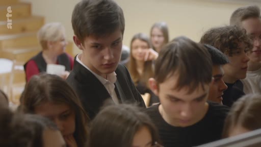 Un groupe d'élèves, dont un jeune homme au premier plan, est réuni dans une salle de classe lors d'une discussion ou d'un cours. L'ambiance est studieuse et concentrée, typique d'un environnement scolaire.
