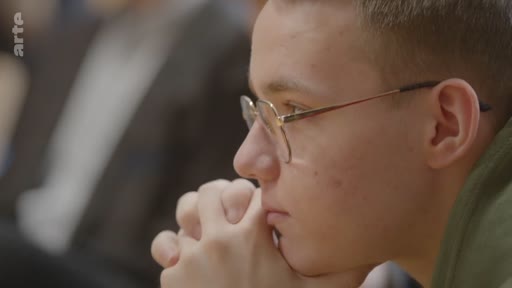 Un jeune étudiant ukrainien portant des lunettes écoute attentivement lors d'une conférence sur l'avenir de l'Ukraine. Il est filmé en gros plan, les mains jointes sous le menton, dans un cadre académique ou de débat.