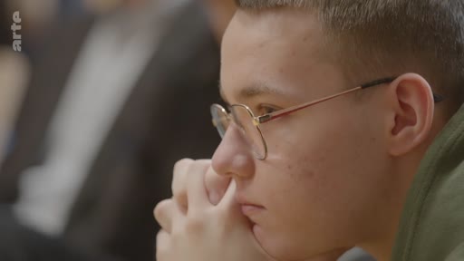 Un jeune étudiant ukrainien, portant des lunettes, écoute attentivement lors d'une discussion sur l'avenir de l'Ukraine. Il est filmé en gros plan, les mains jointes, dans un cadre académique ou de conférence.