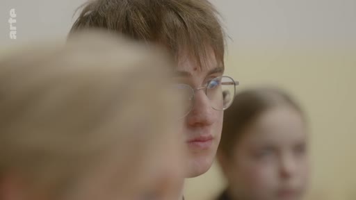 Un jeune homme aux cheveux bruns et portant des lunettes est filmé en gros plan lors d'une séquence de réflexion ou d'entretien. Il est entouré d'autres personnes, suggérant un cadre scolaire ou une réunion de groupe.