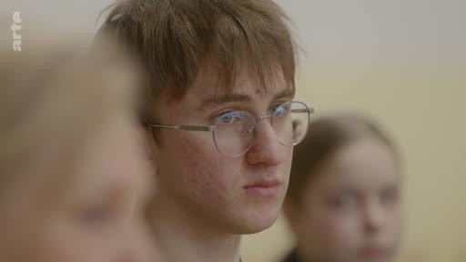 Un jeune homme aux cheveux bruns et portant des lunettes est filmé en gros plan, exprimant une réflexion personnelle ou une attention soutenue lors d'un échange.