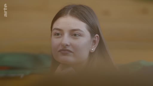 Une jeune femme au regard pensif, filmée en gros plan, semble écouter une intervention lors d'un échange ou d'une réunion.
