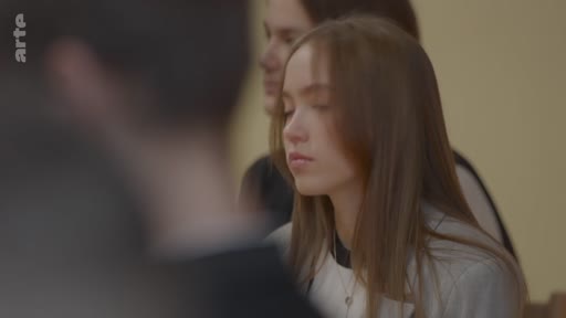 Une jeune femme aux cheveux longs est filmée en gros plan dans une salle, avec une autre personne partiellement visible en arrière-plan. L'ambiance suggère un cadre scolaire ou une réunion formelle.