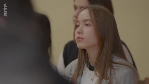 Une jeune femme aux cheveux longs et châtains écoute attentivement une personne hors champ lors d'une discussion en intérieur. Elle semble participer à un échange ou une réunion.