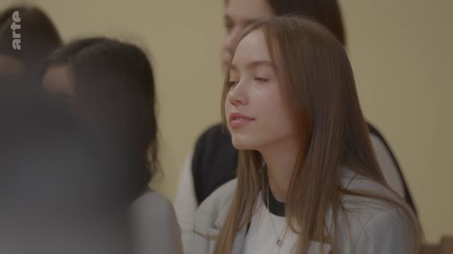 Une jeune femme aux cheveux longs et châtains est filmée en gros plan, semblant écouter avec attention lors d'un événement en intérieur, probablement une salle de classe ou une conférence.
