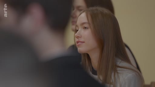 Une jeune femme aux cheveux longs est filmée en gros plan alors qu'elle écoute une conversation dans un cadre scolaire ou professionnel.
