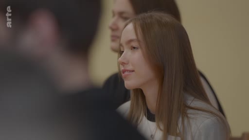 Une jeune femme aux cheveux longs et châtains sourit pendant une interaction sociale ou une réunion, entourée d'autres personnes partiellement visibles.