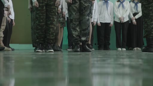 Des élèves, certains en treillis militaires et d'autres en chemises blanches avec des foulards, participent à un exercice de drill dans une école. La scène illustre une formation paramilitaire ou une activité patriotique au sein d'un établissement scolaire.