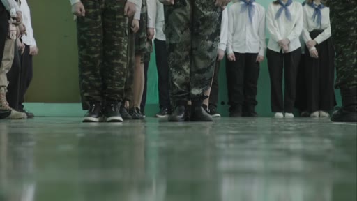 Des élèves, certains en tenue de camouflage et d'autres en chemise blanche avec un foulard bleu, sont alignés lors d'un exercice militaire dans une école. La scène montre une formation disciplinée dans un cadre scolaire.