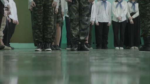 Des élèves participent à un exercice de drill militaire dans une école, portant des uniformes de camouflage et des tenues de cérémonie avec des foulards bleus. La scène illustre l'éducation patriotique ou militaire au sein d'un établissement scolaire.