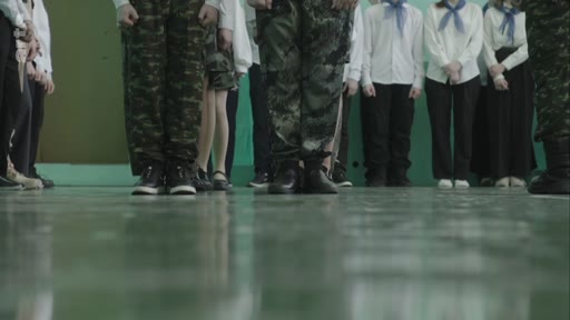 Des élèves participent à un exercice de drill militaire dans une école, portant des uniformes de camouflage et des tenues scolaires traditionnelles avec des foulards. La scène illustre l'éducation patriotique ou militaire au sein d'un établissement scolaire.