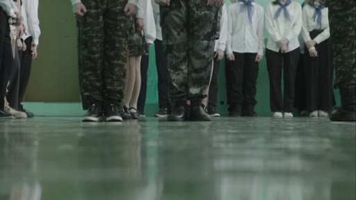 Des élèves, certains en uniforme militaire et d'autres en tenue scolaire avec des foulards, participent à un exercice de drill dans une salle de classe ou un gymnase. La scène illustre une formation militaire ou patriotique au sein d'un établissement scolaire.
