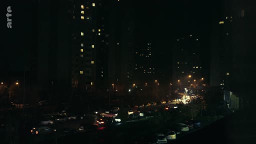 Vue nocturne d'un quartier urbain dense avec des immeubles d'habitation éclairés et une rue où circulent des véhicules, illustrant le contexte d'une zone résidentielle en soirée.