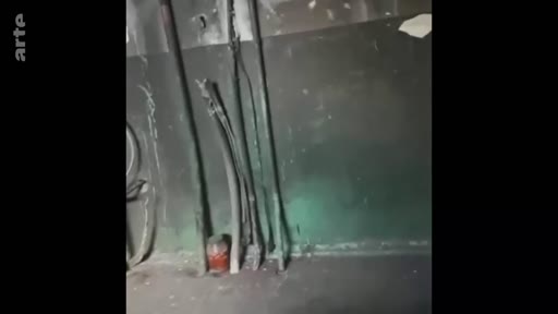 Vue intérieure d'un appartement marqué par les stigmates de la guerre, montrant des tuyaux métalliques endommagés contre un mur sombre et dégradé. Cette image illustre les dégâts matériels et la destruction causés par un conflit armé dans une zone résidentielle.