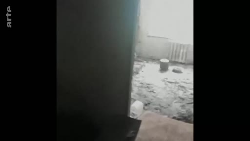 Vue intérieure d'une pièce en ruines, probablement dans une zone de conflit, montrant des débris au sol et un radiateur sous une fenêtre. L'image témoigne des destructions matérielles causées par la guerre.