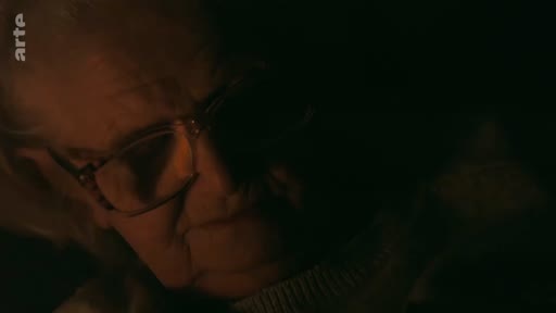 Gros plan sur le visage d'une personne âgée portant des lunettes, filmée dans une ambiance sombre et intimiste, évoquant un témoignage ou une archive personnelle.