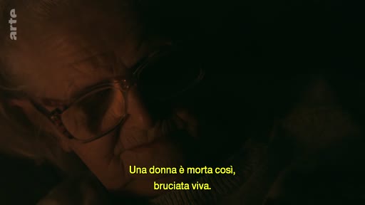 Une personne âgée s'exprime lors d'un témoignage filmé dans l'obscurité, évoquant le décès tragique d'une femme brûlée vive.