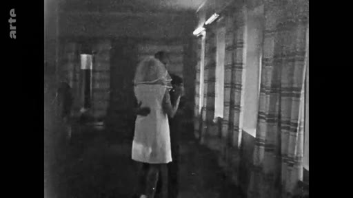 Une image d'archive en noir et blanc montrant un couple en train de danser, la femme porte une robe courte et un voile de mariée, dans une salle intérieure avec des rideaux.