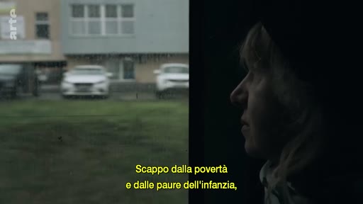 Une femme observe le paysage défilant par la fenêtre d'un véhicule, illustrant un voyage vers une nouvelle vie. Le sous-titre italien évoque la fuite de la pauvreté et des peurs de l'enfance.