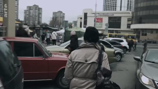 Vue de rue urbaine avec des voitures garées et des passants marchant sur le trottoir, dans un environnement urbain dense avec des immeubles en arrière-plan.