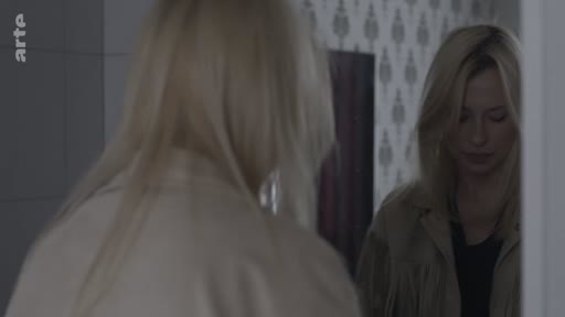 Une jeune femme blonde se regarde dans un miroir, se préparant pour une sortie ou un rendez-vous. Elle porte une veste beige et semble pensive dans cet intérieur domestique.