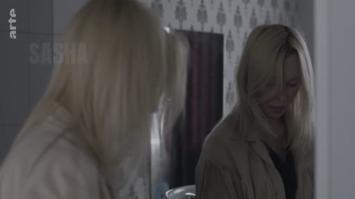 Une femme blonde se regarde dans un miroir, se préparant pour une sortie dans le cadre de la série Sasha.