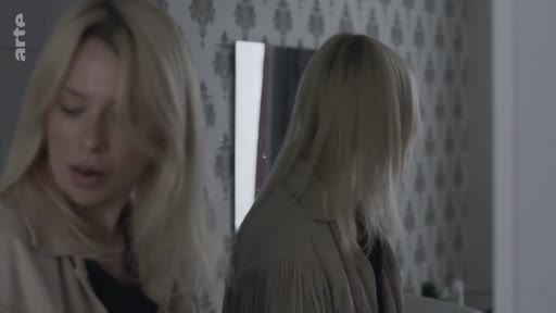 Une femme blonde, vue de face et de dos via son reflet dans un miroir, se prépare dans une pièce au papier peint à motifs.