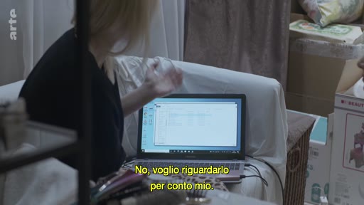 Une femme blonde est assise devant un ordinateur portable ouvert, visionnant des fichiers numériques dans une pièce remplie de cartons et d'objets personnels. Le sous-titre indique : « No, voglio riguardarlo per conto mio ».
