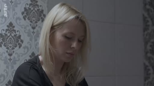 Une femme blonde, filmée en gros plan, semble plongée dans ses pensées ou dans une réflexion mélancolique dans un intérieur avec un papier peint à motifs.