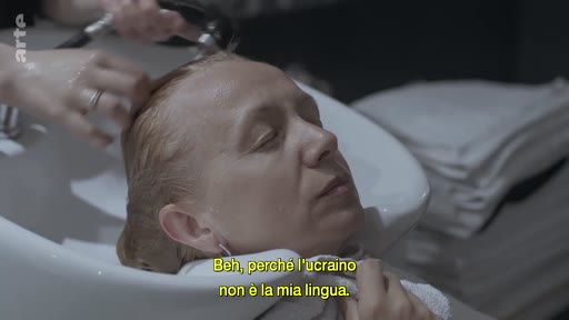 Une femme se fait laver les cheveux dans un salon de coiffure, accompagnée d'un sous-titre en italien évoquant la langue ukrainienne. Cette scène illustre un moment de soin personnel dans un contexte de réflexion sur l'identité et la langue.