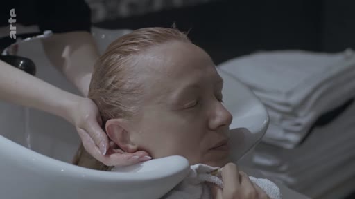 Une femme aux yeux fermés se fait laver les cheveux par une coiffeuse dans un salon de coiffure, dans une scène évoquant un moment de détente ou de soin personnel.