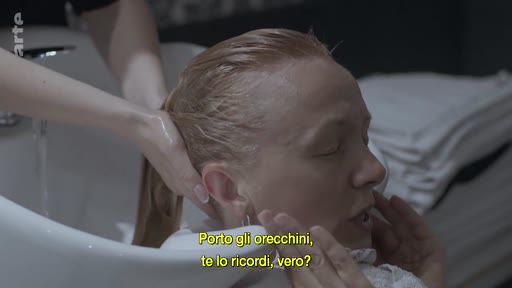 Une femme blonde se fait laver les cheveux dans un bac de lavage de salon de coiffure, tout en discutant. Le sous-titre italien indique : 'Porto gli orecchini, te lo ricordi, vero?'