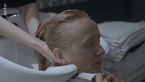 Une femme blonde se fait laver les cheveux par une coiffeuse dans un salon de coiffure, dans une scène évoquant le soin personnel ou un moment de détente.