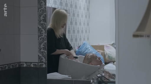 Une femme blonde se fait laver les cheveux par une coiffeuse dans un salon, capturé dans le reflet d'un miroir. Cette scène évoque un moment de soin personnel ou de préparation dans un cadre intime.