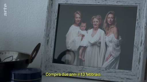 Une photographie de famille encadrée posée sur un meuble, représentant quatre générations de femmes en robes blanches. Le sous-titre indique : « Compirà due anni il 13 febbraio ».